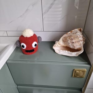 Puede incluir: Una figura roja de ganchillo con una parte superior blanca y orejas grises se encuentra junto a una gran concha. La figura tiene ojos negros y una boca negra. Los objetos est&aacute;n sobre un mueble verde con un asa dorada.