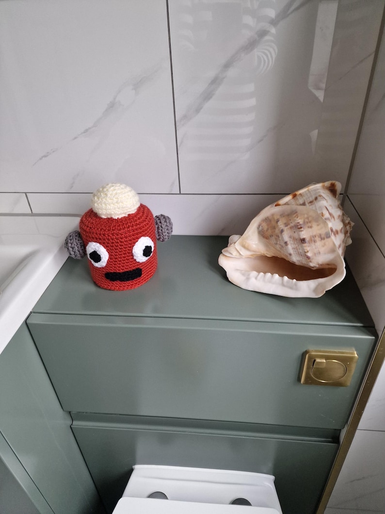 Puede incluir: Una figura roja de ganchillo con una parte superior blanca y orejas grises se encuentra junto a una gran concha. La figura tiene ojos negros y una boca negra. Los objetos est&aacute;n sobre un mueble verde con un asa dorada.