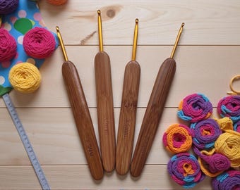 May include: Four wooden crochet hooks with gold-coloured metal tips are arranged on a light wood surface. The hooks are accompanied by colourful yarn balls and small yarn flowers in shades of pink, yellow, orange, and blue, suggesting a crafting theme.