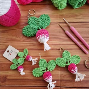 May include: Handmade crochet radish-themed accessories, including earrings and a keychain, are displayed on a wooden surface. The earrings and keychain feature green leaves and pink and white radishes. Yarn and crochet hooks are also visible.