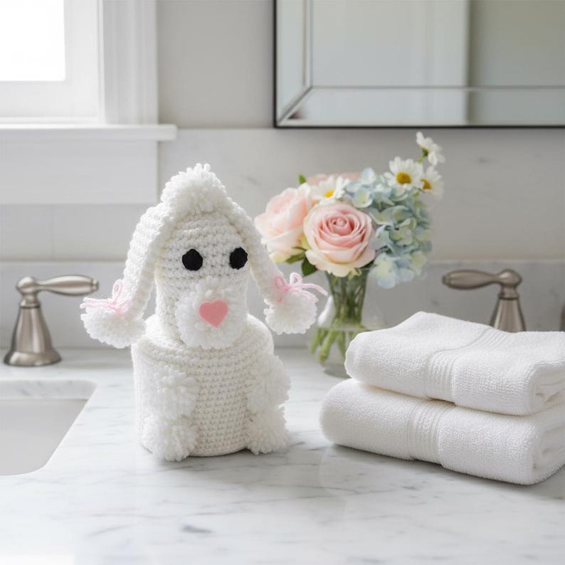 Puede incluir: Un portarrollos de papel higi&eacute;nico con forma de perro tejido a ganchillo blanco con ojos negros y nariz de coraz&oacute;n rosa. El perro tiene orejas largas con detalles rosas y pompones. Dos toallas blancas apiladas.
