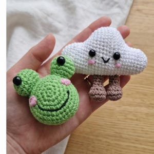 Peut inclure: Deux jouets crochet&eacute;s &agrave; la main : une grenouille verte avec des taches roses et un visage souriant, et un nuage blanc avec un visage souriant et des jambes marron. Les deux jouets ont des yeux en boutons noirs. La grenouille a un corps rond et le nuage est duveteux.