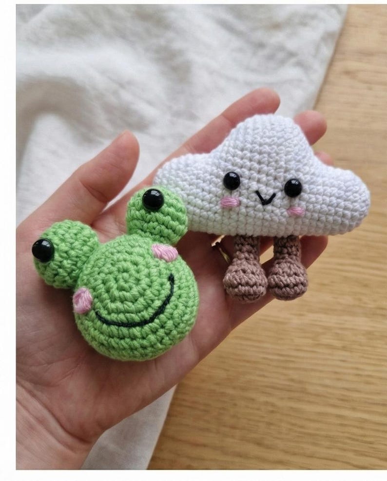 Peut inclure: Deux jouets crochet&eacute;s &agrave; la main : une grenouille verte avec des taches roses et un visage souriant, et un nuage blanc avec un visage souriant et des jambes marron. Les deux jouets ont des yeux en boutons noirs. La grenouille a un corps rond et le nuage est duveteux.