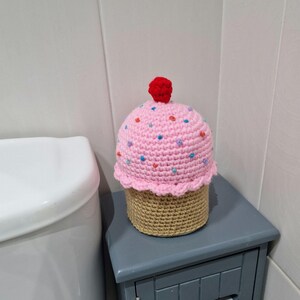 Puede incluir: Una funda de papel higi&eacute;nico con forma de cupcake hecha a ganchillo. La parte superior rosa est&aacute; adornada con chispas de colores y una cereza roja. La base es de color canela y est&aacute; sobre un mueble gris junto a un inodoro blanco.