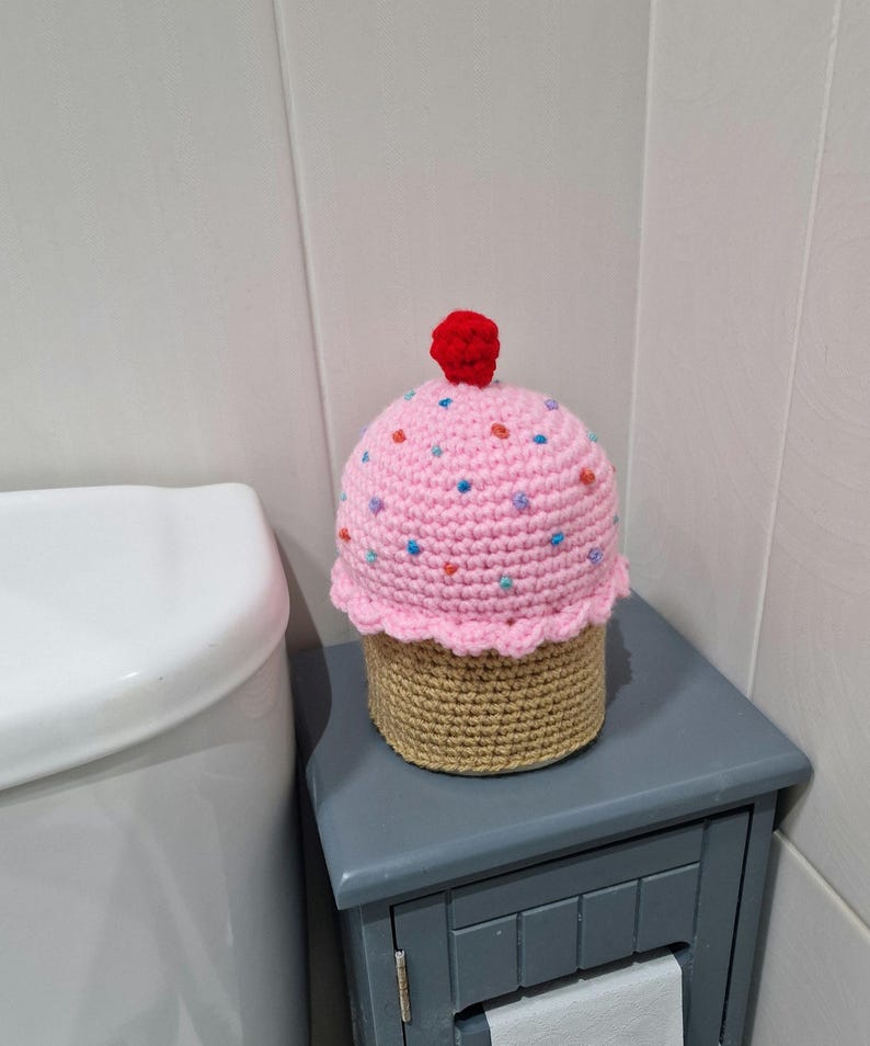 Puede incluir: Una funda de papel higi&eacute;nico con forma de cupcake hecha a ganchillo. La parte superior rosa est&aacute; adornada con chispas de colores y una cereza roja. La base es de color canela y est&aacute; sobre un mueble gris junto a un inodoro blanco.