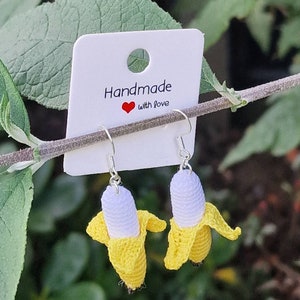 Crochet banana earrings.  These handmade dangle earrings feature small, yellow and white crocheted bananas hanging from silver ear wires.  The earrings are displayed on a white card with the text 'Handmade with love'. The background is blurred greenery.