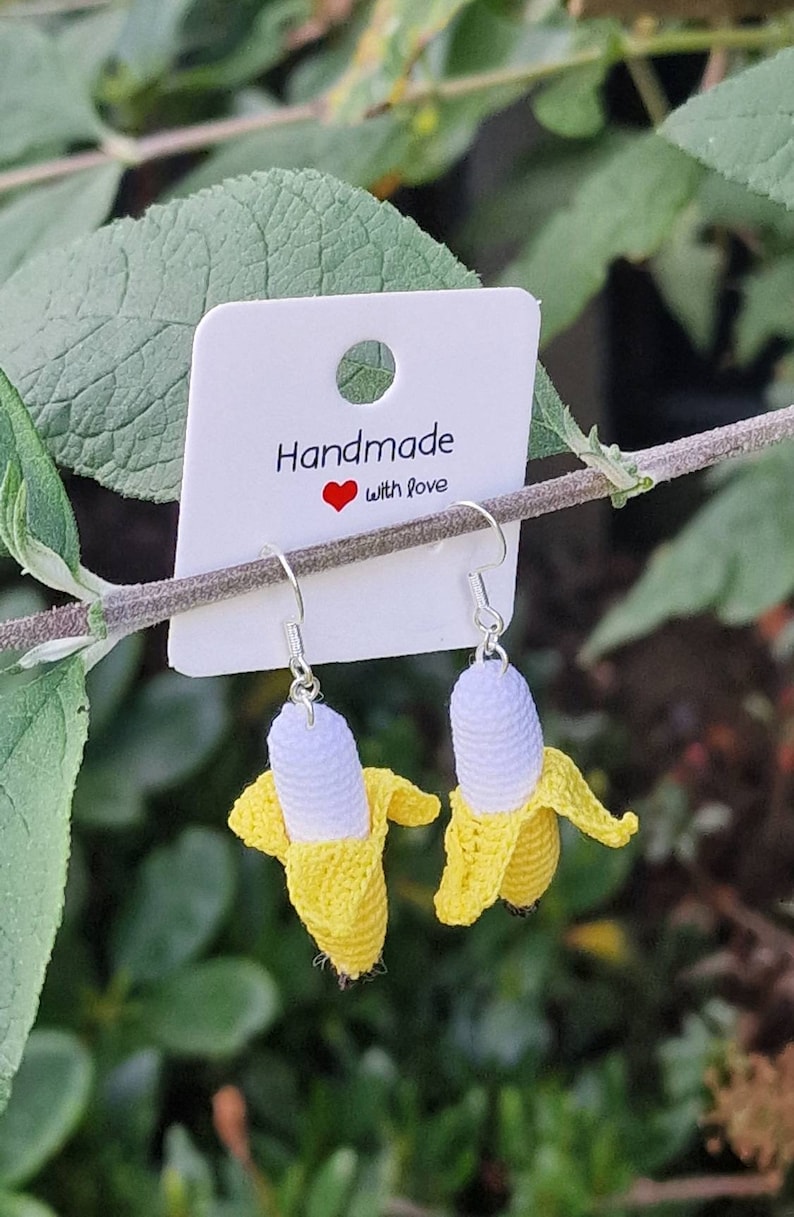Crochet banana earrings.  These handmade dangle earrings feature small, yellow and white crocheted bananas hanging from silver ear wires.  The earrings are displayed on a white card with the text 'Handmade with love'. The background is blurred greenery.