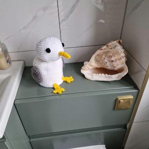 Puede incluir: Un gaviota de ganchillo blanca con alas grises y pico y patas amarillas, posada sobre un mueble verde. Junto a ella, una gran concha. El fondo es un ba&ntilde;o con una pared de azulejos blancos y grises.