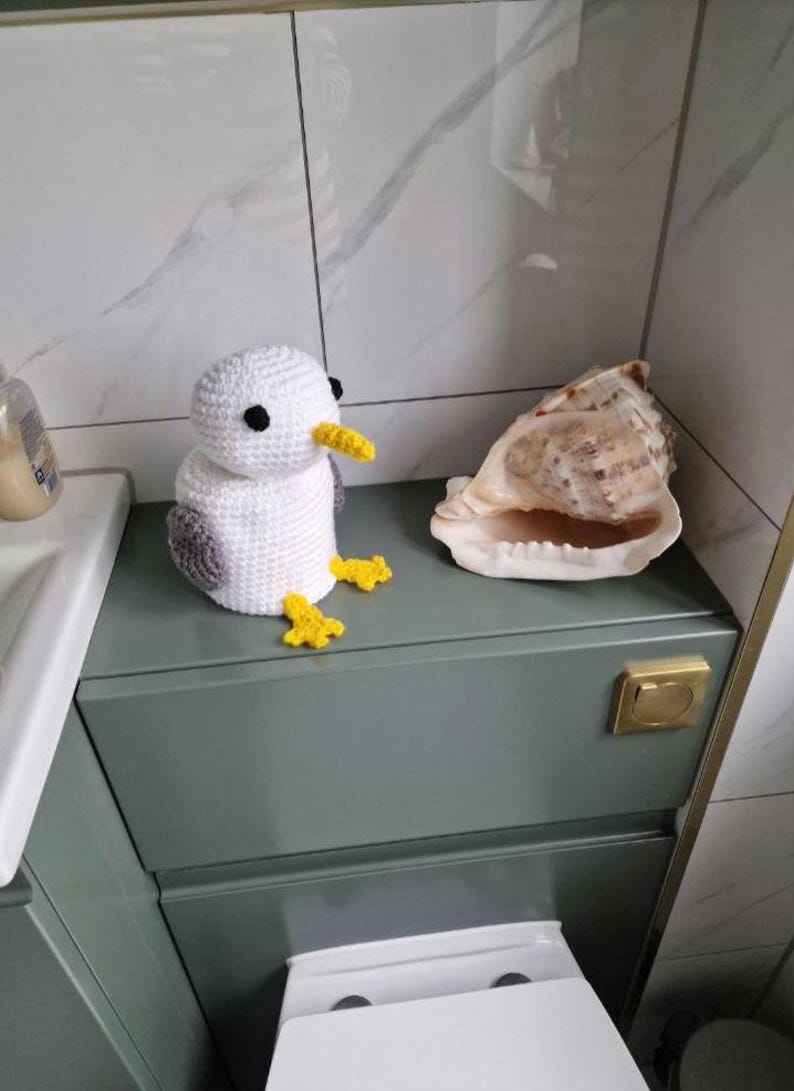 Puede incluir: Un gaviota de ganchillo blanca con alas grises y pico y patas amarillas, posada sobre un mueble verde. Junto a ella, una gran concha. El fondo es un ba&ntilde;o con una pared de azulejos blancos y grises.