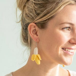 May include: Crocheted banana earrings. The earrings feature a yellow banana peel with a white banana interior. The earrings are suspended from silver-colored hooks. The banana earrings are a fun and unique accessory.