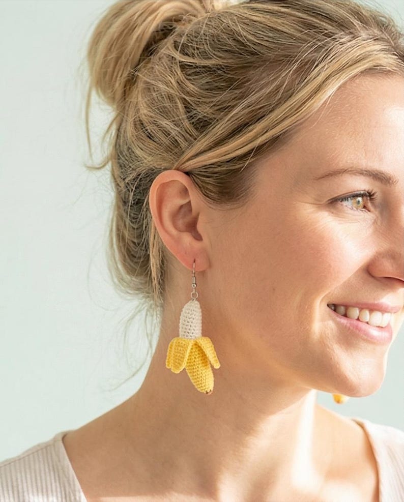 May include: Crocheted banana earrings. The earrings feature a yellow banana peel with a white banana interior. The earrings are suspended from silver-colored hooks. The banana earrings are a fun and unique accessory.