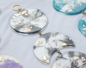 May include: A set of five resin ornaments with gold leaf accents. The ornaments are in the shape of circles and crescent moons. The circles are black and white with gold leaf accents. The crescent moons are black and white, purple and white, and blue and white with gold leaf accents.