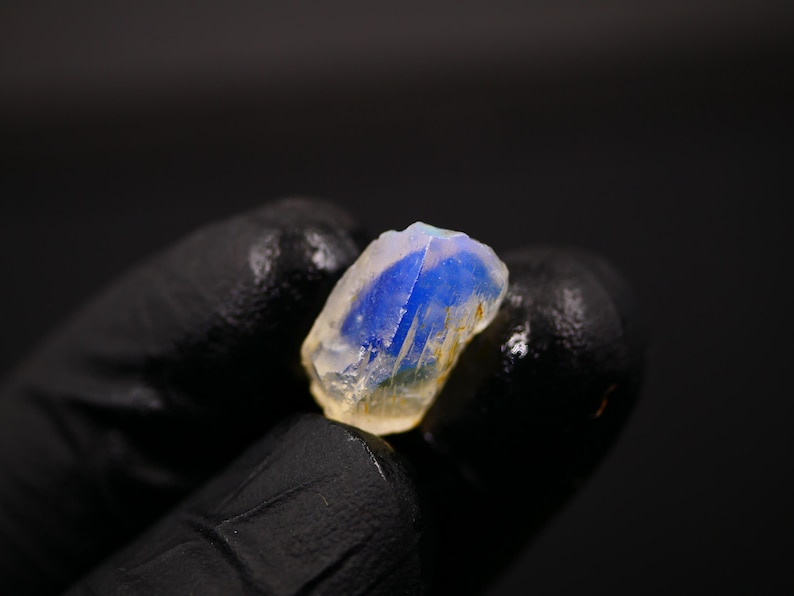 May include: A raw, uncut gemstone with a striking blue interior and a translucent, off-white exterior. The stone is held against a dark background, highlighting its natural form and color variations. The gem's rough texture and unique color make it a captivating mineral specimen.