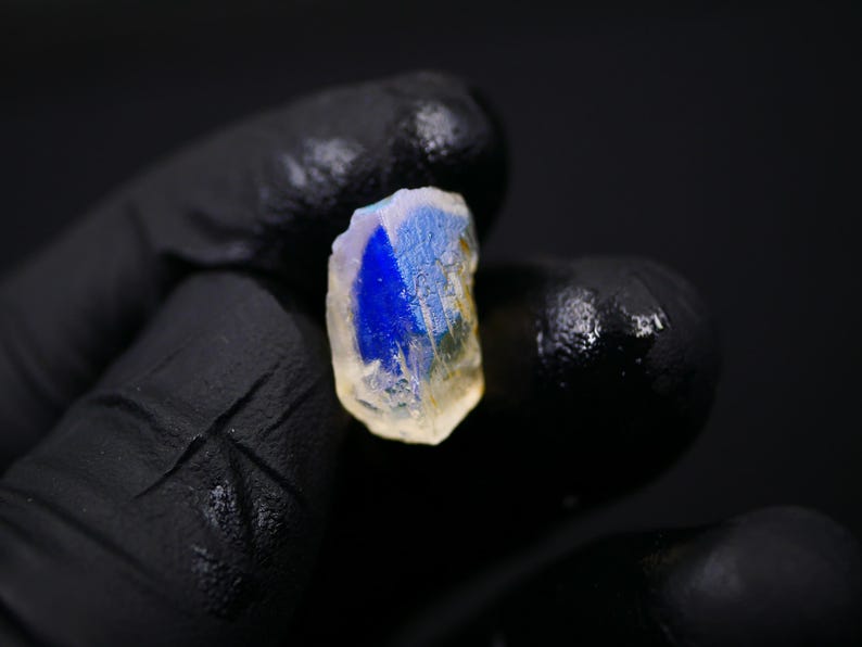 May include: A close-up of a gemstone with a vibrant blue and yellow color. The gemstone is held in a black gloved hand against a black background. The stone has a rough, natural texture.