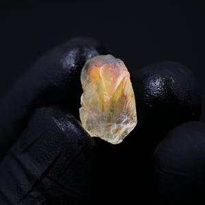 May include: A rough, translucent gemstone with a mix of yellow, orange, and hints of green. The stone is held against a black background, showcasing its natural, irregular shape and internal textures. The gemstone appears to be raw and uncut.