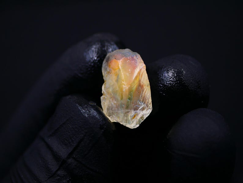 May include: A rough, translucent gemstone with a mix of yellow, orange, and hints of green. The stone is held against a black background, showcasing its natural, irregular shape and internal textures. The gemstone appears to be raw and uncut.