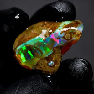 May include: A raw, unpolished opal gemstone featuring a brown, textured matrix with vibrant flashes of neon green, electric blue, and bright pink iridescent color play across its surface, showcasing a natural, irregular, and organic shape.