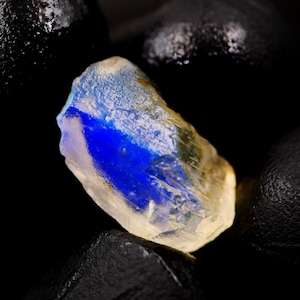 May include: A raw, uncut gemstone with a striking blue and yellow color gradient. The gem's surface is textured, and the blue hue is concentrated in a central band. The stone is set against a dark, contrasting background.