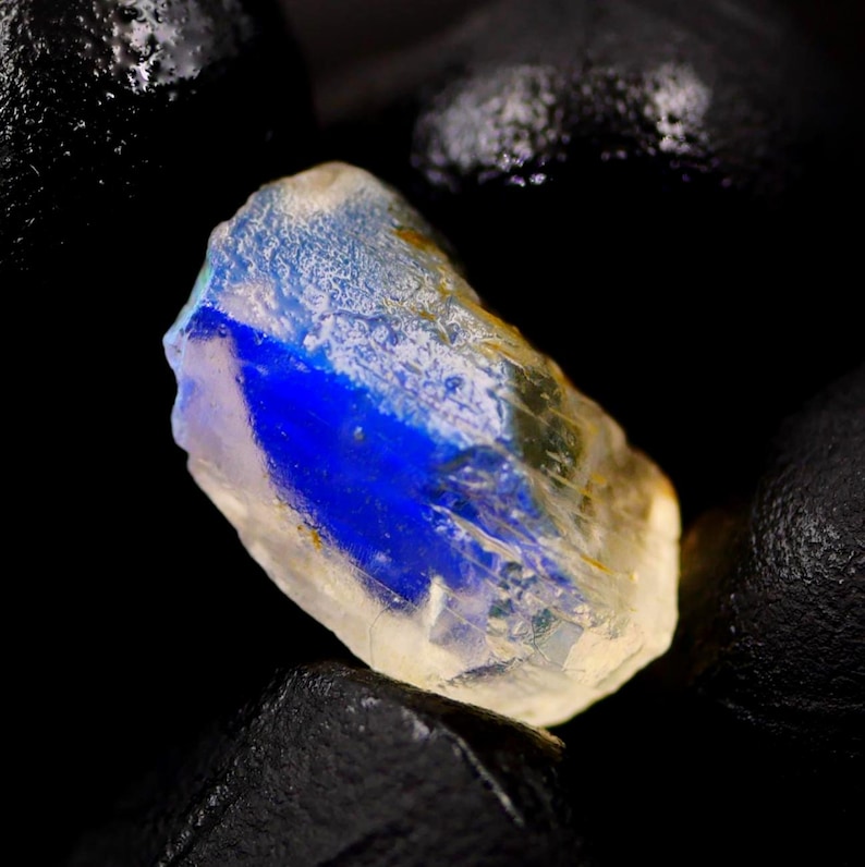 May include: A raw, uncut gemstone with a striking blue and yellow color gradient. The gem's surface is textured, and the blue hue is concentrated in a central band. The stone is set against a dark, contrasting background.