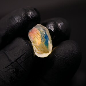 May include: A close-up of a raw, uncut gemstone held in a gloved hand. The stone displays a mix of colors, including blue, yellow, and pink, with a translucent appearance. The background is dark.