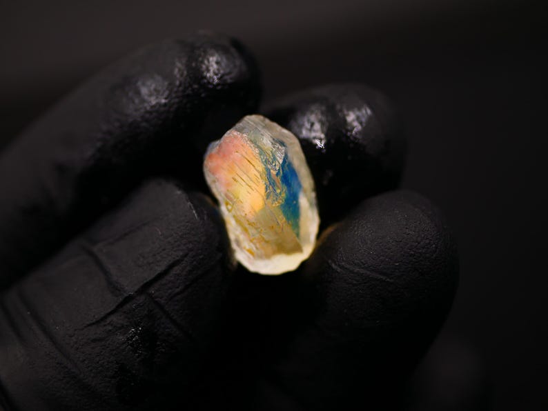 May include: A close-up of a raw, uncut gemstone held in a gloved hand. The stone displays a mix of colors, including blue, yellow, and pink, with a translucent appearance. The background is dark.
