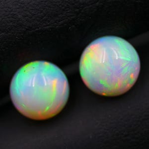 May include: Two round opals with iridescent colors of green, orange, and yellow. The opals are white with a glossy finish. The gemstones are set against a dark background.
