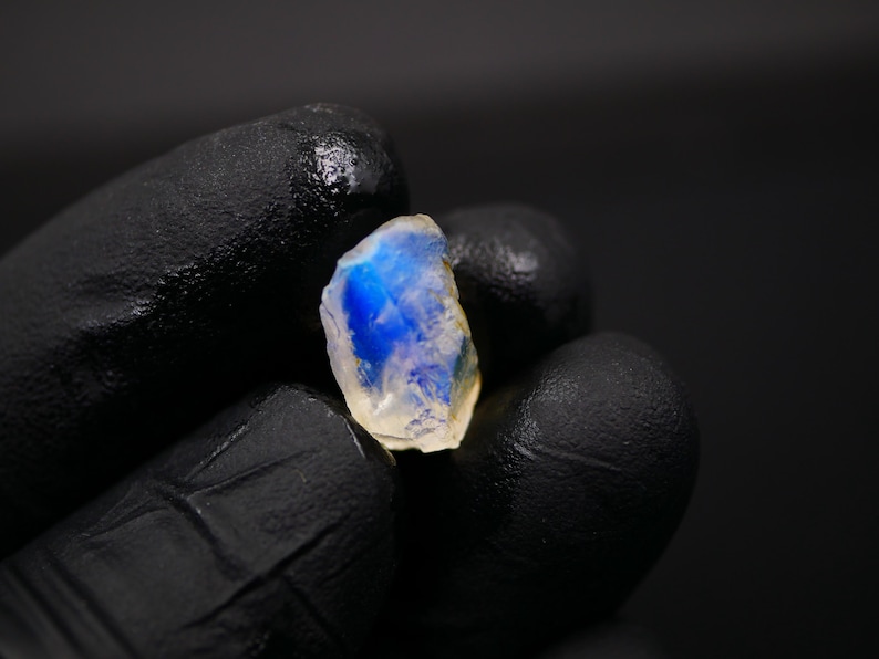 May include: A small, rough-cut gemstone with a translucent, off-white base and a vibrant blue interior. The stone is held against a black background, highlighting its natural texture and color variations. The gem is likely a moonstone.