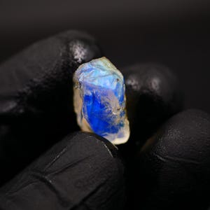 May include: A raw, uncut gemstone with a vibrant blue and white color. The stone is held against a dark background, showcasing its natural, irregular shape and unique color variations. This gemstone is a beautiful example of natural mineral formations.