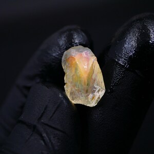 May include: A rough-cut, translucent gemstone with a mix of yellow, orange, and pink hues. The stone is held against a black background, showcasing its natural, irregular shape and crystalline structure. The gem appears to be raw and unpolished.