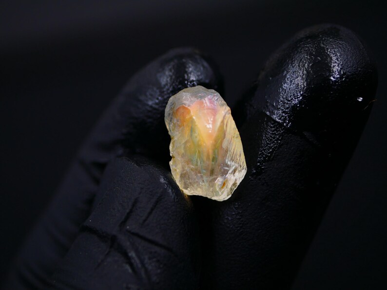 May include: A rough-cut, translucent gemstone with a mix of yellow, orange, and pink hues. The stone is held against a black background, showcasing its natural, irregular shape and crystalline structure. The gem appears to be raw and unpolished.