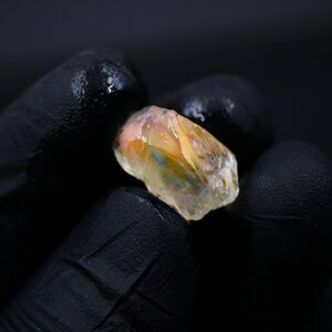May include: A rough-cut gemstone with a mix of colors, including orange, yellow, and blue, is held in a gloved hand. The stone has a translucent quality, and the background is dark.