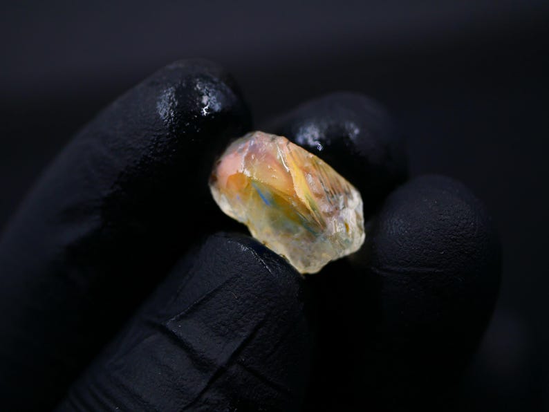 May include: A rough-cut gemstone with a mix of colors, including orange, yellow, and blue, is held in a gloved hand. The stone has a translucent quality, and the background is dark.