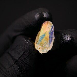 May include: A raw, uncut gemstone with a mix of colors, including yellow, orange, and blue. The stone is held against a black background, highlighting its natural, rough texture and unique color variations. The gemstone is a small, irregular shape.