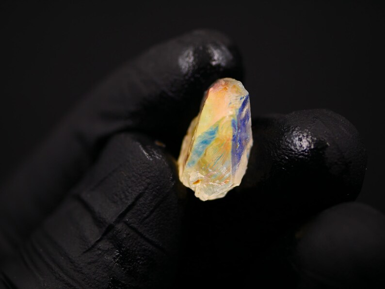 May include: A raw, uncut gemstone with a mix of colors, including yellow, orange, and blue. The stone is held against a black background, highlighting its natural, rough texture and unique color variations. The gemstone is a small, irregular shape.