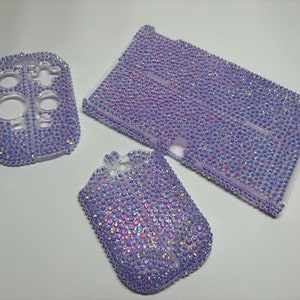 May include: Three purple and iridescent rhinestone-covered cases for a gaming console. The cases are for the console, the controllers, and the game cartridges.