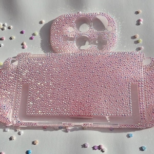 Peut inclure: Un étui et une coque de manette de console de jeu roses, incrustés de strass étincelants. La coque de la manette est placée au-dessus de l'étui de la console. De petites perles colorées sont éparpillées autour de l'étui et de la coque. L'étui mesure environ 15 cm de long.