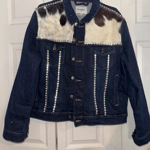 May include: A dark blue denim jacket with a cowhide patch on the front and rhinestone detailing. The jacket has a button closure and long sleeves.