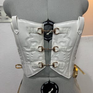 May include: A white corset with a western-inspired design. The corset features decorative stitching, gold-colored hardware, and adjustable straps. The garment is displayed on a mannequin.