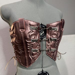 May include: A metallic rose-colored corset with a western-inspired design. The corset features decorative stitching and panels with a faux fur accent. It is secured with metal clasps and laces.