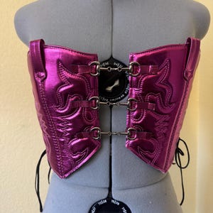 May include: A metallic fuchsia corset, crafted from repurposed cowboy boot material, is displayed on a mannequin. The corset features intricate stitching, silver-tone hardware, and adjustable straps. The design incorporates classic western boot elements.