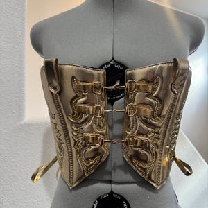 May include: Gold-colored corset with intricate embossed detailing and multiple buckle closures. The corset is displayed on a mannequin, showcasing its shape and design. The corset has decorative stitching and small gold accents.