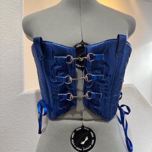 May include: A royal blue corset with decorative stitching and silver-colored metal clasps. The corset is displayed on a mannequin and features adjustable straps and satin ribbon ties. The overall design is a blend of classic and modern styles.