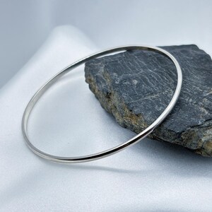 May include: A silver bangle bracelet with a minimalist design. The round bracelet is displayed on a white, silky surface next to a dark grey rock. The bangle is made of a shiny, reflective metal.