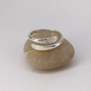 May include: A silver wrap ring with a textured upper section. The ring is presented on a smooth, light brown stone against a white backdrop. The ring's design features an open, adjustable style.