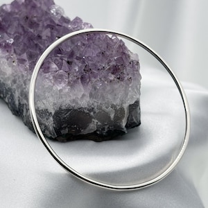 May include: A silver bangle bracelet with a simple, circular design. The bracelet is displayed against a backdrop of a large, purple amethyst crystal and a white, silky fabric.