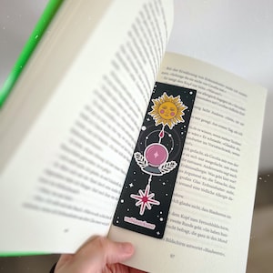 Könnte beinhalten: Ein schwarzer Lesezeichen mit Himmelsillustrationen, darunter eine Sonne, eine Kristallkugel und ein Stern, befindet sich in einem offenen Buch. Der Lesezeichen hat den Text "verblasstekunst". Die Seiten des Buches sind weiß mit schwarzem Text.