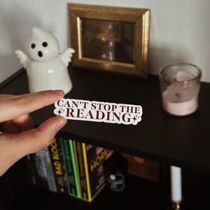 Könnte beinhalten: Ein weißer Aufkleber mit dem burgunderroten Text "CAN'T STOP THE READING". Der Aufkleber wird vor einem schwarzen Bücherregal mit Büchern, einer Geisterfigur, einem gerahmten Bild und einer Kerze gehalten.