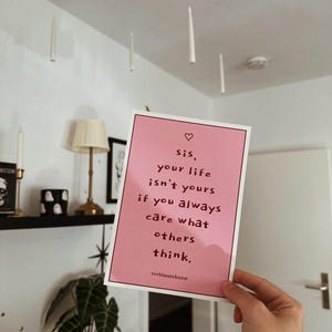 Könnte beinhalten: Ein rosa Druck mit dem Text "sis, your life isn't yours if you always care what others think." Der Druck wird vor einer weißen Wand mit schwebenden Kerzen und einem Regal mit Dekoration gehalten.