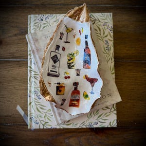 May include: A white oyster shell dish with gold accents, decorated with a colorful illustration of various cocktails, bottles of liquor, and fruit. The dish is sitting on top of a book with a floral design.