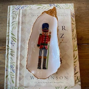 Peut inclure: Un plat en céramique blanche en forme de coquille d'huître avec un bord doré, représentant un casse-noisette de Noël traditionnel peint à la main en rouge et bleu.